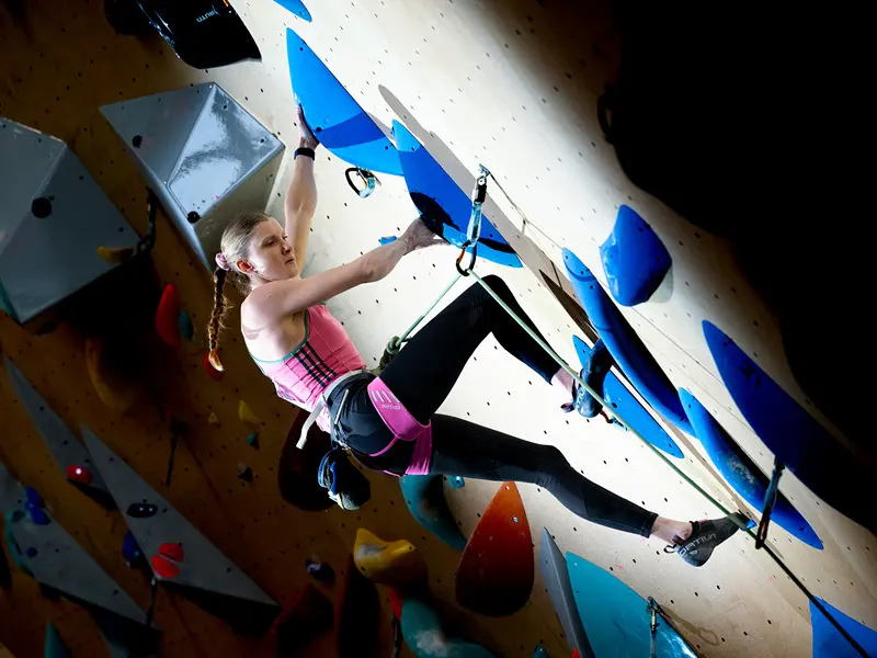 Een vrouw in sportkleding beklimt een indoor-klimwand, terwijl ze zich vastklampt aan blauwe grepen en een klimgordel draagt, en het licht van bovenaf op haar schijnt.